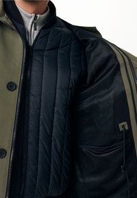 Olivgrön jacka med en svart vadderad foder, med dragkedja fram, meshinredning och en dold ficka för förvaring.