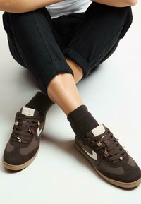 Brązowe zamszowe sneakersy z białym logo, ozdobione charmsami na sznurowadłach, w zestawieniu z prążkowanymi brązowymi skarpetami i podwiniętymi czarnymi spodniami.
