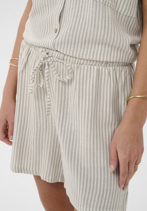 Beige-weiße Shorts mit vertikalen Streifen und Tunnelzug, getragen mit passendem, geknöpftem Oberteil und goldenen Armbändern an den Seiten der Hände.