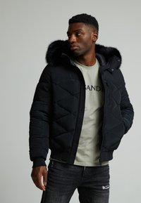 Giacca a vento nera imbottita con cappuccio foderato in pelliccia, design trapuntato, chiusura frontale con zip e polsini aderenti, abbinata a una camicia verde chiaro e jeans scuri.