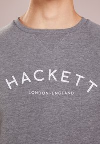 Sudadera gris con cuello redondo y un ligero corte en V. Presenta un gran diseño de texto en blanco que dice "HACKETT LONDON. ENGLAND."