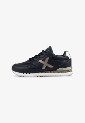 Zapatilla deportiva negra con parte superior de cuero texturizado, acentos de malla gris, un logotipo X marrón y una entresuela moteada. Suela blanca.