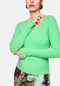 Grüner strukturierter Langarmpullover mit figurnaher Passform, Rundhalsausschnitt und gerippten Bündchen. Kombiniert mit gemusterten Blumenhosen.