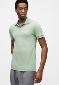 Homme portant un polo vert clair à manches courtes avec col et un pantalon gris, regardant vers sa gauche sur un fond clair uni.