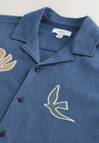 Chemise bleue à boutons avec col, présentant des motifs d'oiseaux et de feuilles brodés sur le devant, et une étiquette de marque « NEXT » à l'intérieur du col.