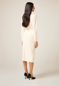 Abito color crema con maniche lunghe decorato con borchie. L'abito ha una vestibilità aderente e una lunghezza a metà polpaccio. Scarpe nere con tacco.