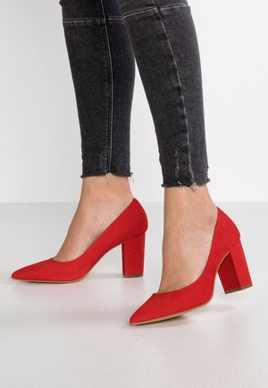 Røde ruskind højhælede sko med spids tå og en kraftig blokhæl, præsenteret sammen med slidte sorte denimjeans.