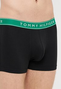 Cuecas boxer pretas de algodão com uma cintura elástica verde apresentando o nome da marca "TOMMY HILFIGER" em letras brancas.
