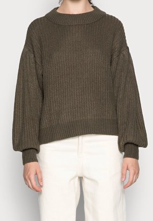 Person wearing a loose, chunky knit olive green sweater with balloon sleeves and ribbed cuffs, paired with high-waisted off-white pants.