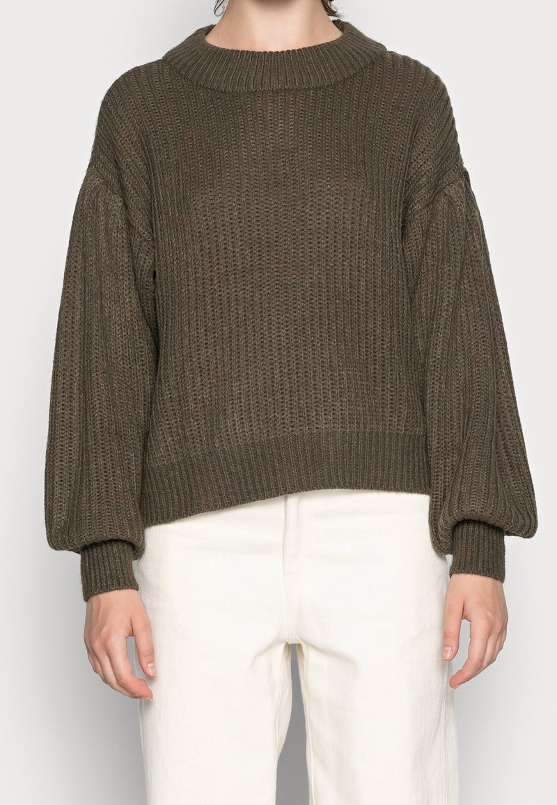 Personne portant un pull large en grosse maille vert olive avec des manches ballon et des poignets côtelés, associé à un pantalon taille haute écru.