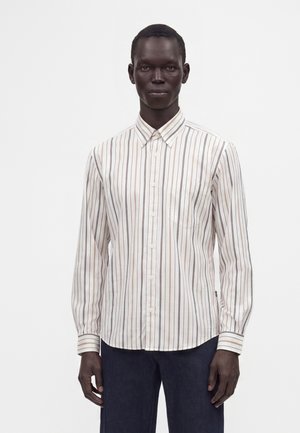 Persona que lleva una camisa blanca de manga larga con botones, con finas rayas verticales marrones y negras y pantalones oscuros, de pie sobre un fondo liso.