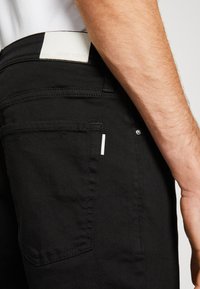 Jeans de denim negro con un parche de logo blanco en la cintura, costuras prominentes y un diseño minimalista con un pequeño bolsillo adicional.