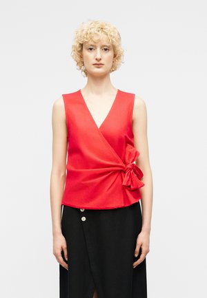 Γυναίκα με ξανθά κατσαρά μαλλιά, που φοράει ένα κόκκινο αμάνικο τοπ με δέσιμο στη μέση και μια μαύρη φούστα με κουμπιά.