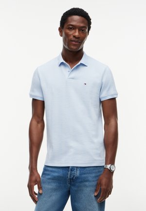 Mann trägt ein hellblaues kurzärmeliges Poloshirt mit kleinem Logo auf der Brust und blaue Jeans, steht vor einem schlichten weißen Hintergrund.