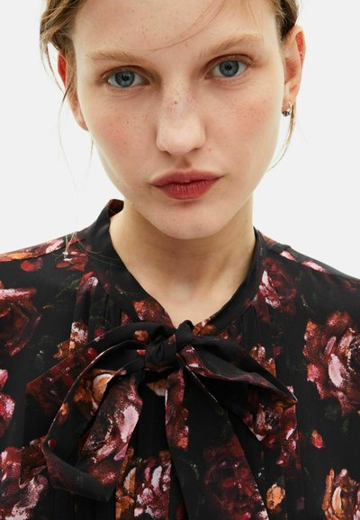 Schwarze Bluse mit floralem Muster in Rot und Pink, mit einem Bindegürtel am Hals und subtilen Falten. Weicher Stoff mit glatter Textur.