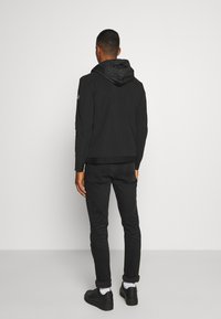 Veste noire à capuche avec une capuche texturée, design ajusté et poignets réglables, associée à un jean noir et des baskets noires.