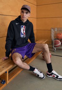 Hanorac negru Nike cu logo-ul NBA, pantaloni scurți de baschet purpurii, șosete negre Nike și pantofi Nike negri/albi/roșii; așezat pe o bancă din lemn.