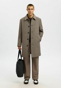 Cappotto herringbone marrone con colletto e bottoni, abbinato a pantaloni beige e una borsa scura; il modello indossa scarpe bianche e nere.