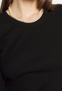 Maglietta nera a costine con maniche lunghe e scollatura tonda, abbinata a una collana di perle bianche che poggia sulla clavicola.
