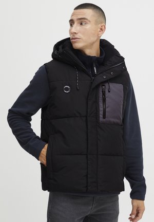 Zwarte puffer vest met capuchon, voorzien van een met rits sluitend borstzakje in donkergrijs. Inclusief patches en een rits aan de voorkant. De textuur is glad.