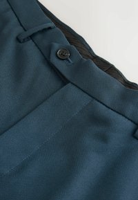 Tmavě tyrkysové kalhoty vyrobené z texturované látky, s černým knoflíkem, poutky na opasek a boční kapsou. Čistý, dobře střižený design.