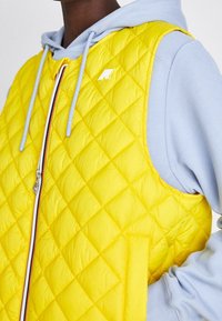 Persona con un hoodie azzurro sotto un gilet giallo trapuntato con una cerniera multicolore e un piccolo logo bianco sul petto.