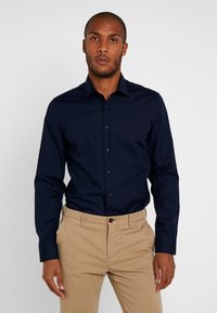 Granatowa koszula zapinana na guziki z gładkiego materiału, z długimi rękawami i standardowym kołnierzykiem, noszona z beżowymi spodniami.