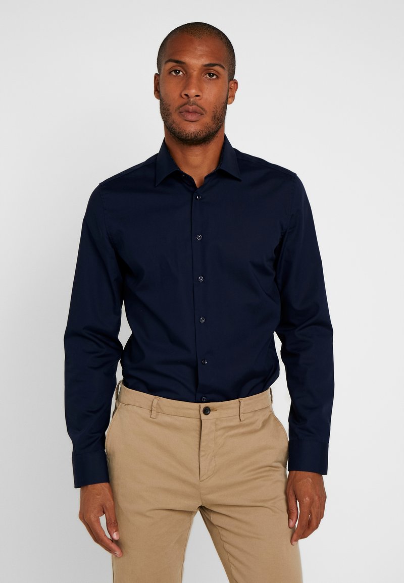 Granatowa koszula zapinana na guziki z gładkiego materiału, z długimi rękawami i standardowym kołnierzykiem, noszona z beżowymi spodniami.