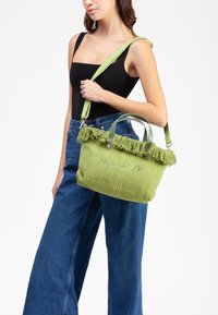 Borsa a tracolla verde in maglia con bordo superiore frangiato, doppi manici e tracolla rimovibile. Presenta il testo ricamato "Plage del Rio."