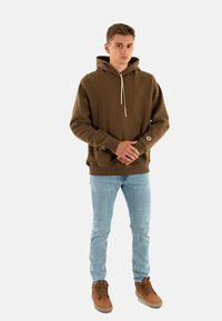 Felpa cappuccio marrone con tasca a marsupio frontale, cordini beige e logo sul polsino, abbinata a jeans azzurri chiari e scarpe marroni.