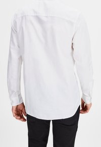Camicia bianca a maniche lunghe con colletto botton-down, taglio dritto e texture liscia, abbinata a pantaloni neri.