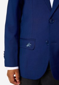 Tumši zils bleizeris ar gludu tekstūru, ar ieliktu kabatu un mazu izšūtu logotipu kabatas malā. Redzami divi pogas.