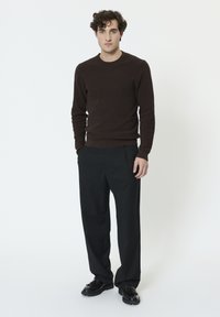 Pull en tricot marron, pantalon noir à jambes larges, et chaussures noires. Le mannequin pose devant un fond blanc, avec des lignes simples et épurées.