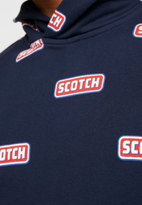 Suéter navy con un alto cuello que presenta parches del logo "SCOTCH" en rojo y blanco en repetición. La tela tiene una textura suave y elástica.
