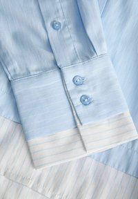Poignets de chemise bleu clair avec deux boutons assortis, motif rayé blanc et bleu subtil sur le tissu.