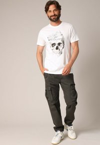 T-shirt blanc avec un motif de skull et du texte. Pantalons cargo gris avec plusieurs poches. Baskets blanches avec des accents bleus.