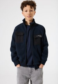 Chaqueta de forro polar azul marino con dos bolsillos negros en el pecho, cierre con cremallera y puños acanalados. Llevada sobre una camisa azul marino y unos jeans grises.