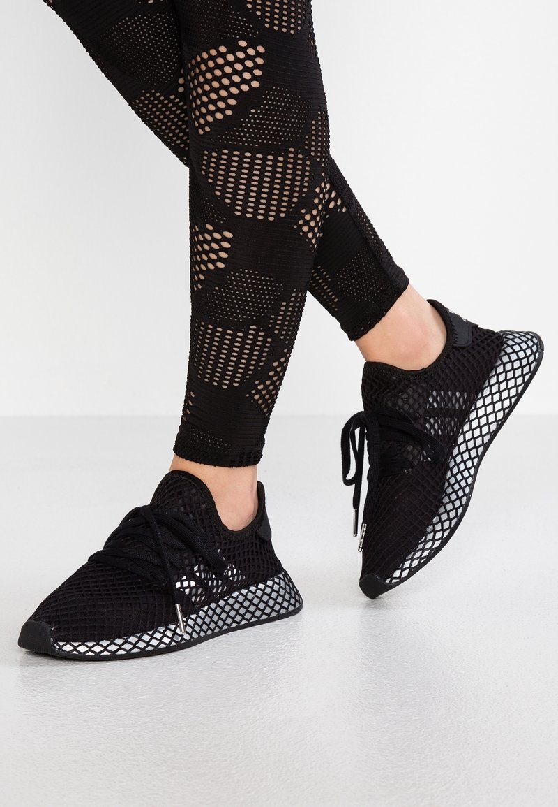 Czarne sneakersy z przewiewną siateczką na cholewce, z teksturowaną powierzchnią, posiadające odbijającą światło podeszwę z wzorem w kratkę oraz sznurowane zapięcie, zestawione z czarnymi legginsami z siateczki.