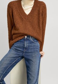 Maglione spesso di colore marrone con scollo a V, abbinato a jeans blu a vita alta. Texture morbida e vestibilità comoda.