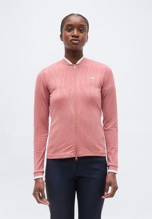 Vrouw met een nauwsluitend, roze, langarm jasje met ritssluiting en subtiele golvende patronen, gecombineerd met een donkere broek, staand tegen een effen achtergrond.