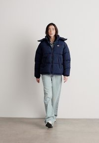 Ναυτικό φούτερ μπουφάν με φερμουάρ μπροστά, κουκούλα και δύο πλαινές τσέπες. Φορεμένο με ανοιχτό μπλε τζιν και μαύρα sneakers σε έναν μινιμαλιστικό χώρο.