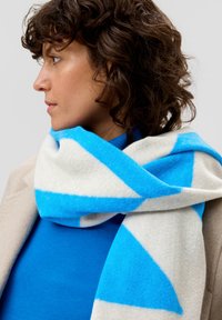 Wollen sjaal in een mix van materialen, met grote blauwe geometrische patronen op een crèmekleurige achtergrond, met een zachte textuur en elegant om de nek gedrapeerd.