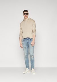 Beige Kapuzenpullover mit Fronttasche, kombiniert mit hellblauen, verwaschenen Skinny Jeans und weißen Sneakers. Modell trägt schwarze Sonnenbrille.