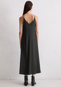 Langes, dunkelgrünes Slipkleid mit einem V-Rücken und dünnen Trägern; glattes, glänzendes Material; kombiniert mit schwarzen Ankle-Boots.