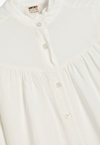 Blusa blanca de botones con diseño plisado, cuello corto y botones transparentes. Textura suave y tejido ligero.