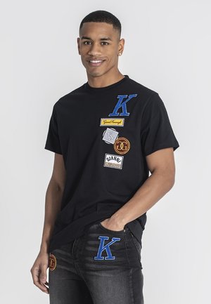 Camiseta negra de algodón con varios parches de tela, incluyendo una letra "K" azul y otros logotipos, combinada con jeans oscuros que muestran acentos similares.
