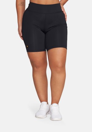 Kobieta w czarnych sportowych szortach z wysokim stanem i boczną kieszenią oraz białych sneakersach, stojąca na jednolitym tle.