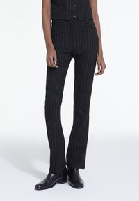 Pantalons noirs à rayures fines en tissu texturé, avec une coupe ajustée et des jambes droites, complétés par des bottes noires à la cheville.