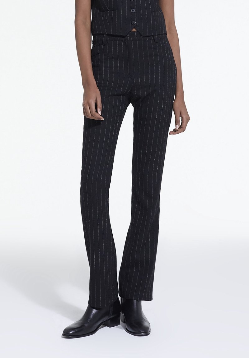 Pantalons noirs à rayures fines en tissu texturé, avec une coupe ajustée et des jambes droites, complétés par des bottes noires à la cheville.