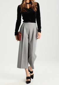 Schwarzes Samt-Oberteil mit langen Ärmeln und durchsichtigen Punkten, kombiniert mit grauen, hochgeschnittenen, weiten Hosen und schwarzen Absätzen. Braune Handtasche inklusive.